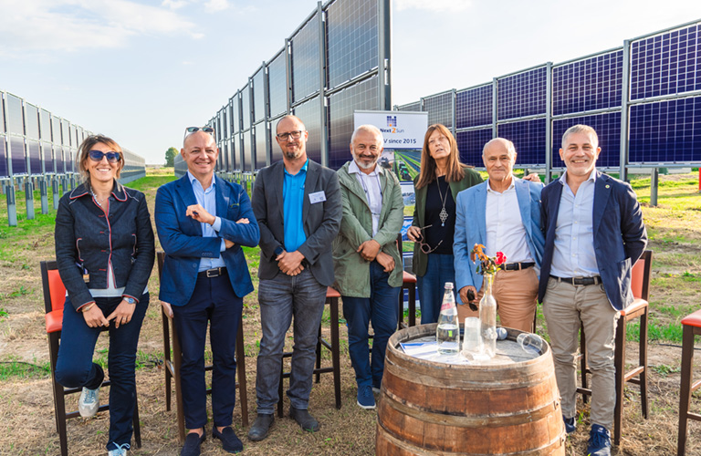 Sieben Personen stehen im Freien zwischen Reihen aufgeständerter Solarmodule eines Agri-Photovoltaik-Parks. Im Vordergrund befindet sich ein runder Stehtisch aus einem Holzfass mit Wasserflaschen und einer kleinen Blumenvase. Im Hintergrund erstrecken sich die Solaranlagen entlang eines Feldes unter hellem Tageslicht. Auf einem Schild zwischen den Modulreihen ist das Logo von Next2Sun sichtbar.
